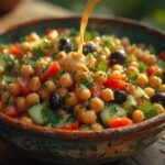 Uma tigela azulada com uma salada de grão-de-bico, pepino, tomate e azeitonas, sendo temperada com molho tahini, destacando o frescor do prato.