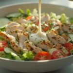 Close-up apetitoso de uma salada de frango grelhado com tiras suculentas de frango sobre alface, tomate cereja e pepino, sendo regada com um molho cremoso de iogurte e hortelã.