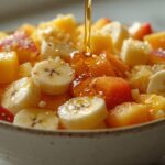 Close-up de uma salada de frutas refrescante com pedaços de manga, banana, mamão e outros, com mel sendo derramado por cima.