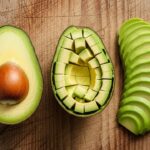 Imagem mostrando como cortar abacate de três formas sobre uma tábua: uma metade com caroço, outra metade picada em cubos na casca e a polpa em fatias.